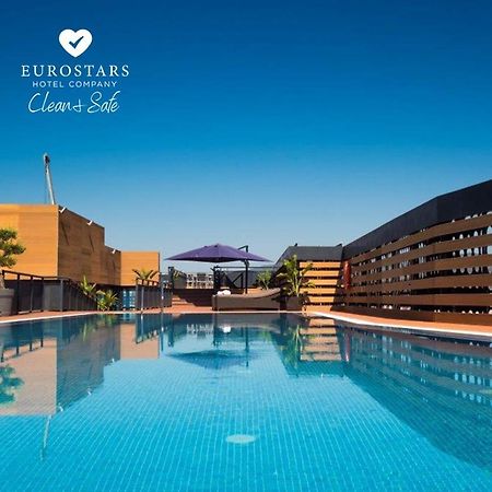 Eurostars Palace Córdoba Exterior foto The photo shows an outdoor swimming pool with clear, blue water, reflecting the blue sky above. The pool area is surrounded by modern architectural features, including wooden slats and potted plants. There is also a purple umbrella providing shade. T