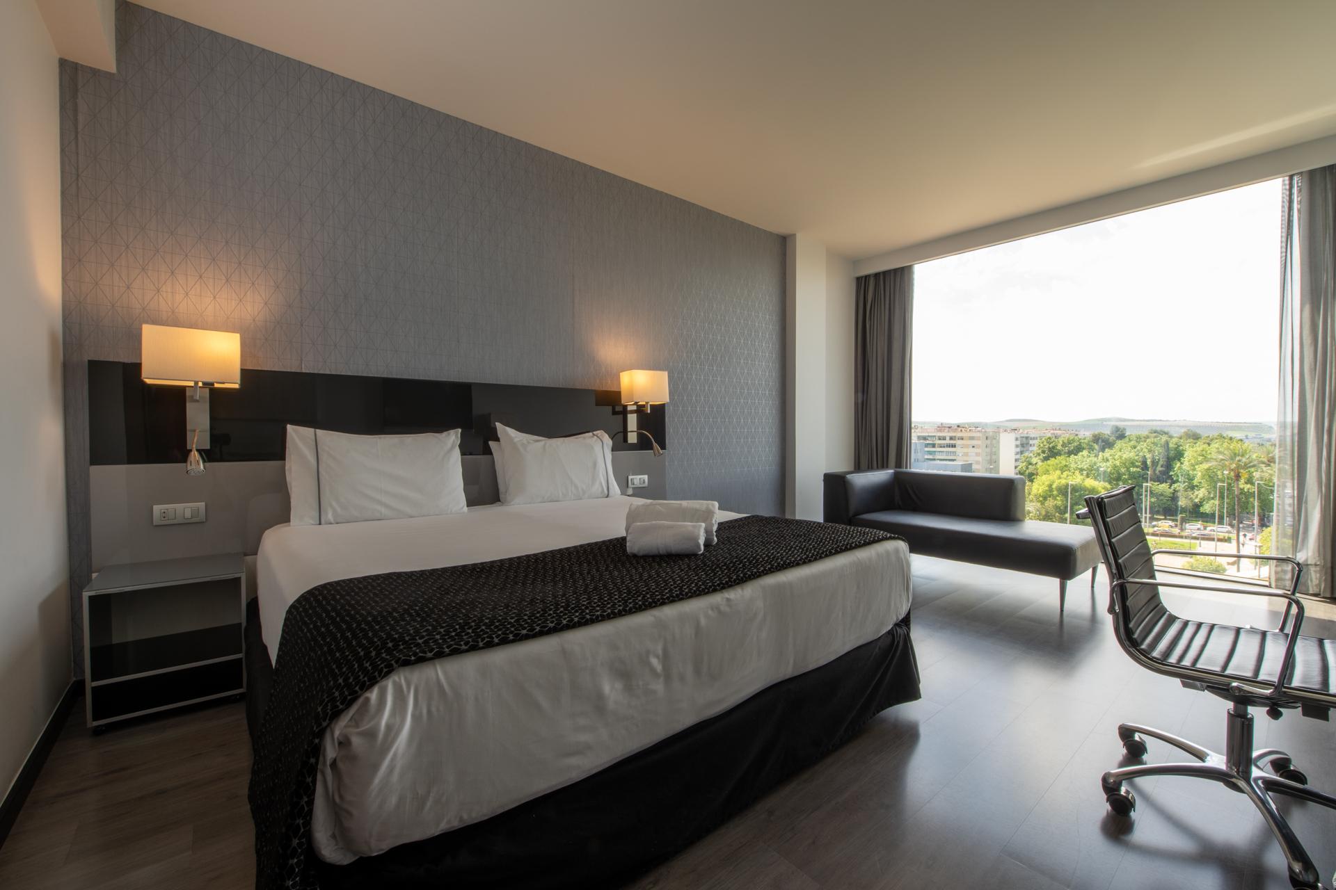 Eurostars Palace Córdoba Exterior foto The photo shows a modern hotel room featuring a large bed with white linens and a dark patterned throw. On either side of the bed, there are stylish wall-mounted lamps. A contemporary desk chair is positioned at a small desk, and there’s a sofa near 