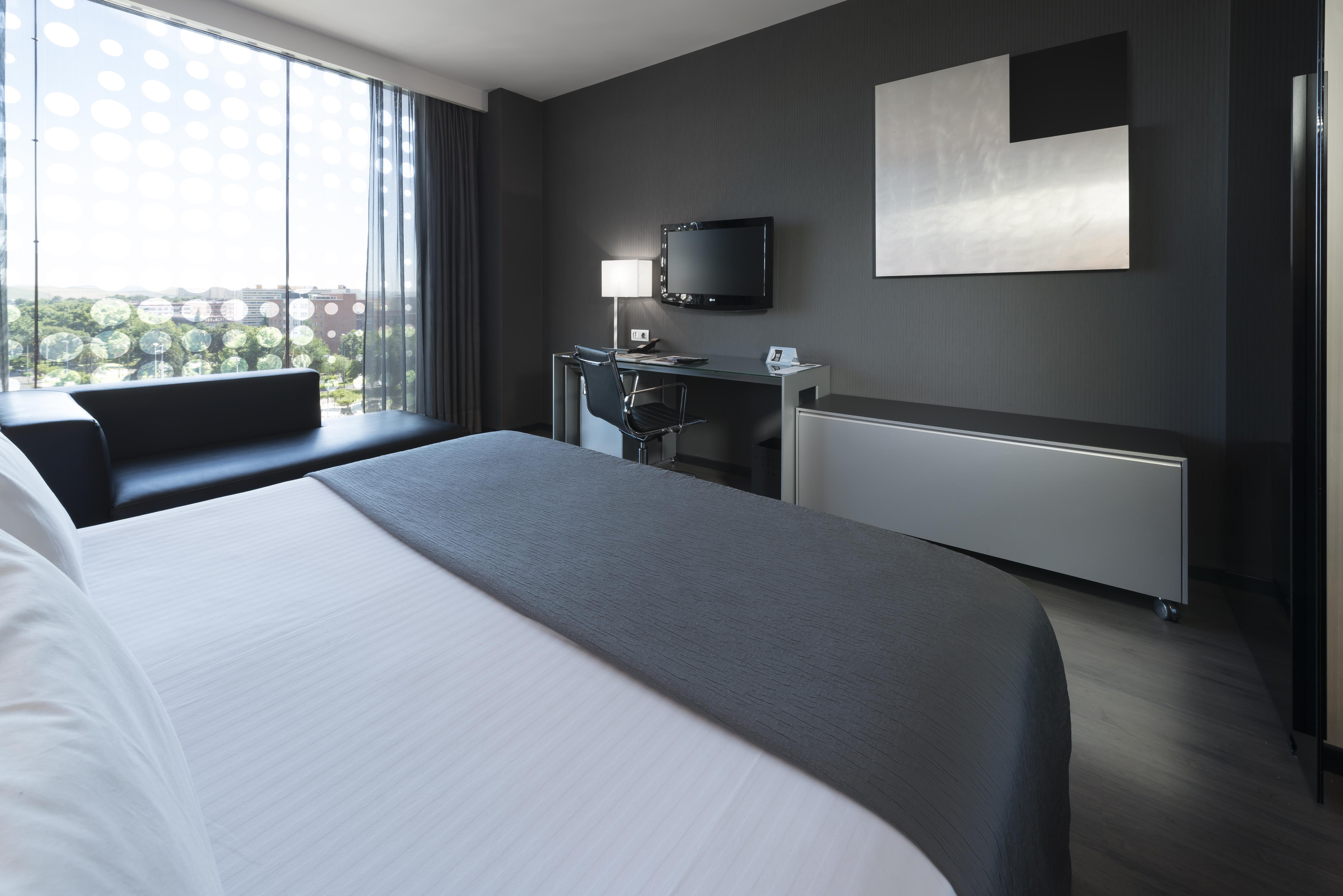 Eurostars Palace Córdoba Exterior foto The photo shows a modern hotel room featuring a sleek design. In the foreground, there is a bed with a white coverlet and gray accents. To the side, a dark sofa is visible. The room has a large window with sheer curtains, allowing natural light to en