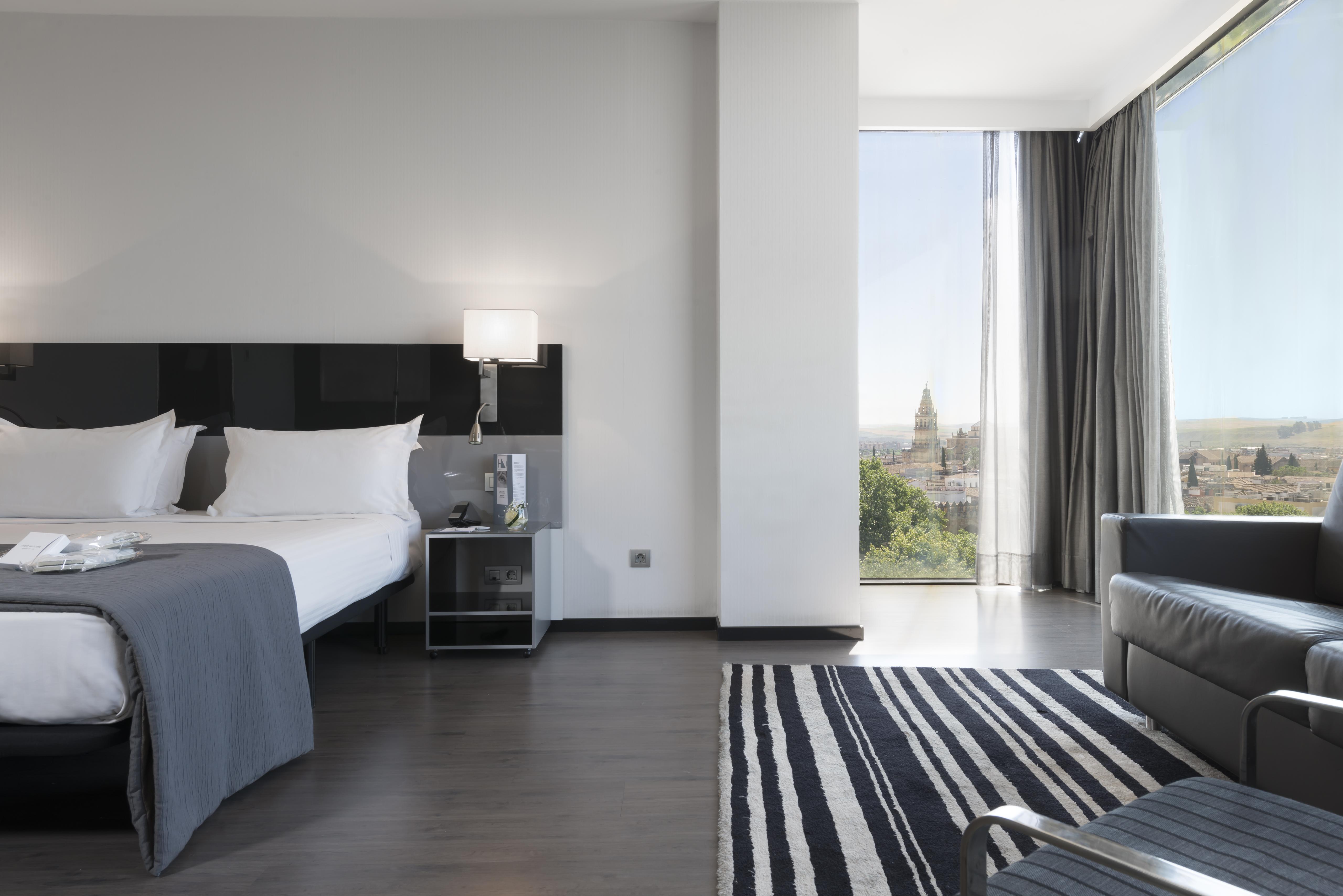 Eurostars Palace Córdoba Exterior foto The photo shows a modern hotel room featuring a large bed with white linens and dark cushions, paired with a sleek nightstand. There is a contemporary chair and a small table nearby, along with a striped area rug on the floor. A large window allows n