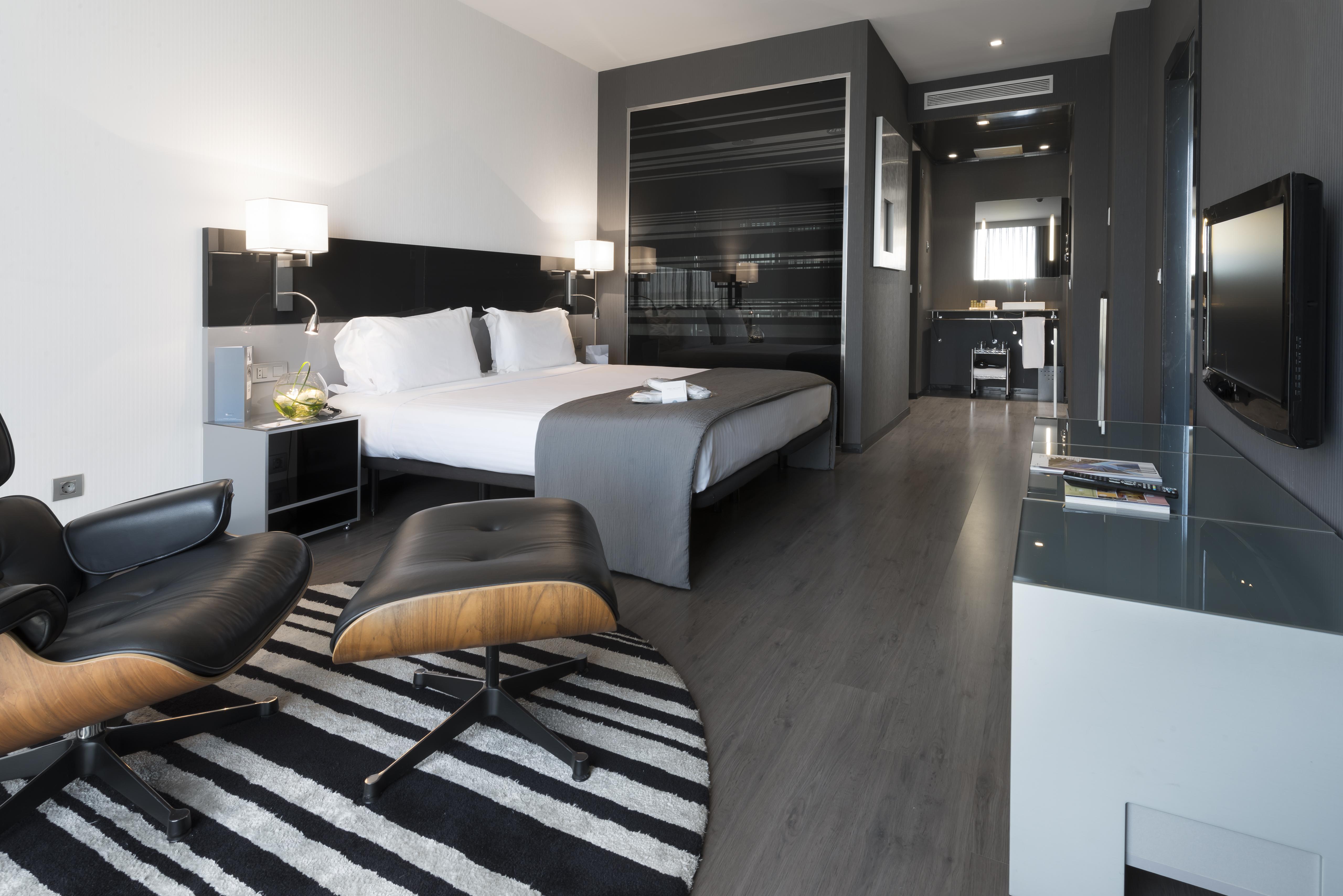 Eurostars Palace Córdoba Exterior foto The photo shows a modern hotel room featuring a king-sized bed with light-colored linens and dark accent walls. There are stylish bedside tables with lamps, and a comfortable lounge chair paired with an ottoman placed on a striped rug. The room also 