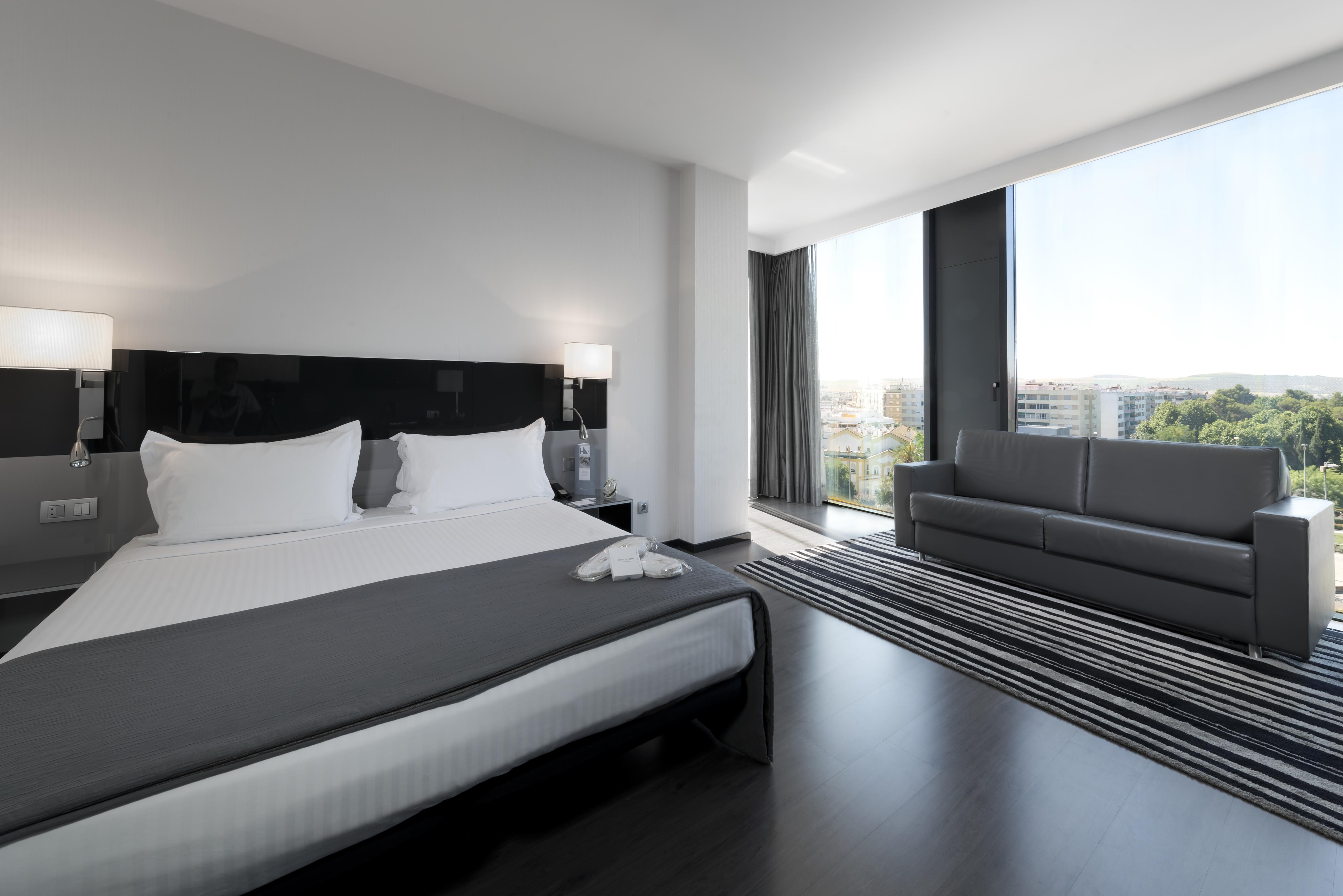 Eurostars Palace Córdoba Exterior foto The photo shows a modern hotel room with a sleek design. It features a large bed with white bedding and two pillows, accompanied by a gray throw. To the side of the bed, there are stylish nightstands with lamps. A large window allows natural light to