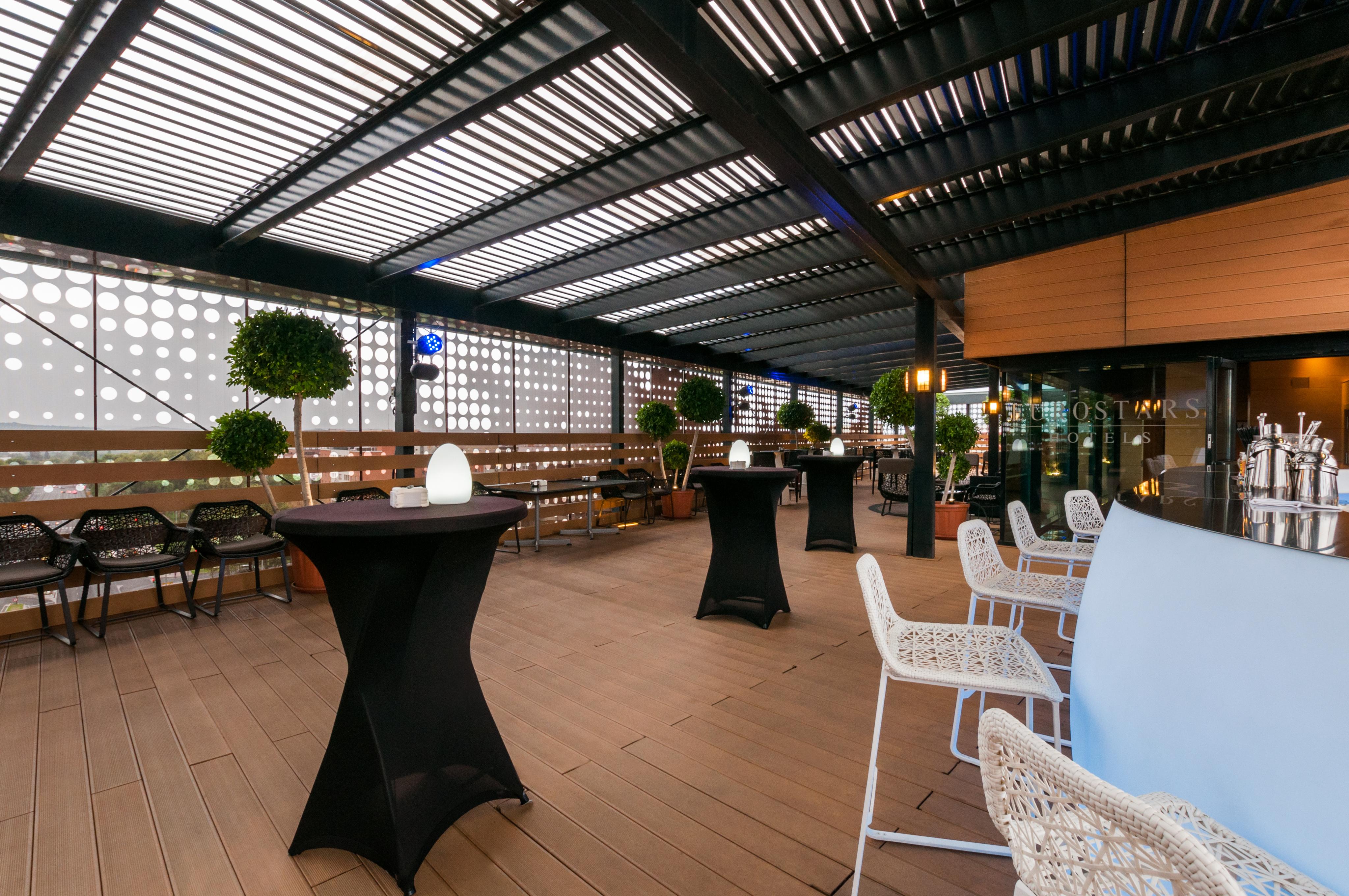 Eurostars Palace Córdoba Exterior foto The photo shows a stylish outdoor seating area, likely part of a restaurant or lounge. It features a wooden floor and an overhead structure with horizontal slats, allowing for both shade and light. There are several tall, black cocktail tables and wh