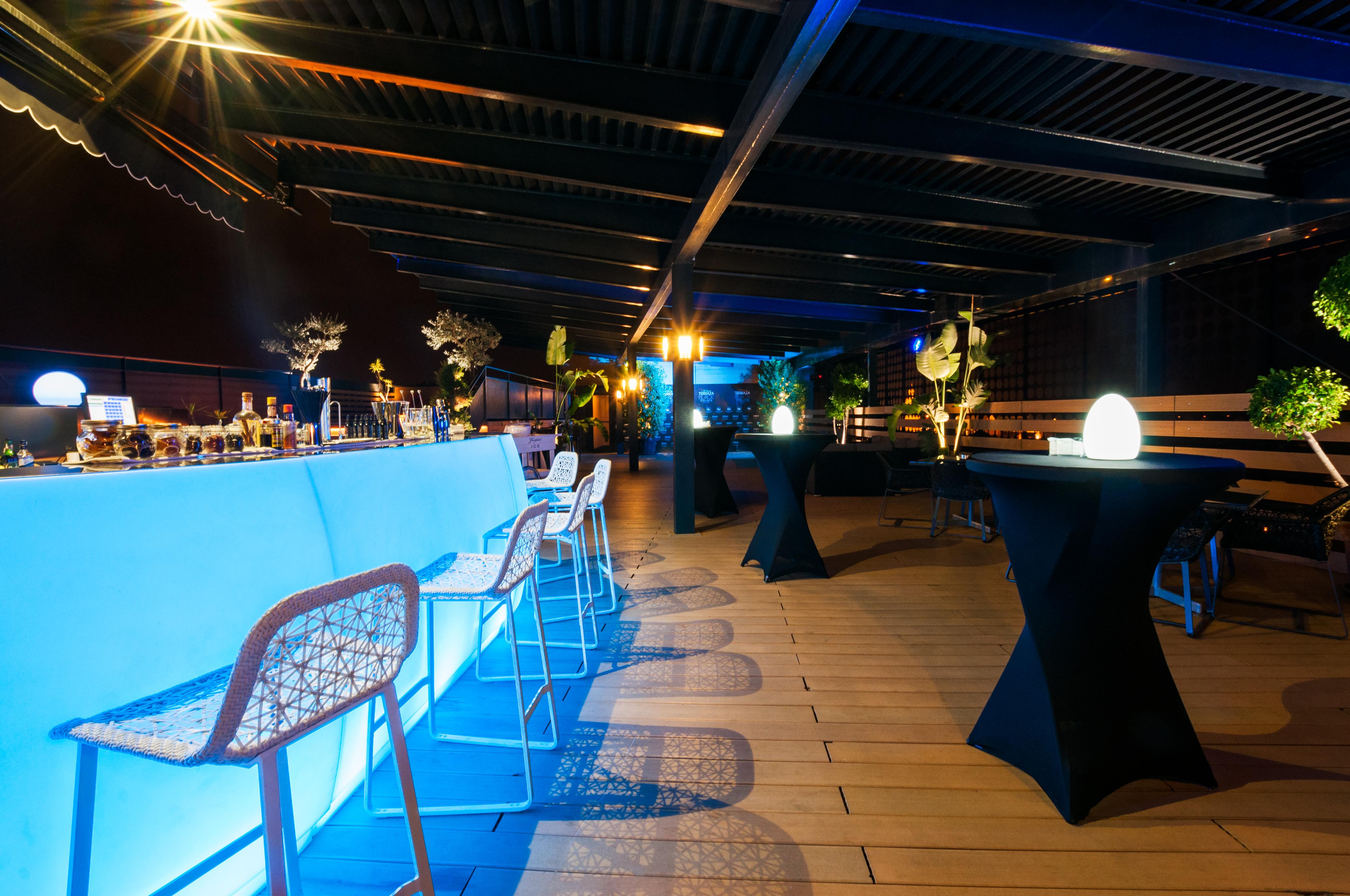 Eurostars Palace Córdoba Exterior foto The photo shows a stylish rooftop or outdoor bar area. There is a long bar counter illuminated with blue lighting, complemented by modern bar stools. The space is decorated with potted plants and elegant round tables covered in black cloth. Soft ligh