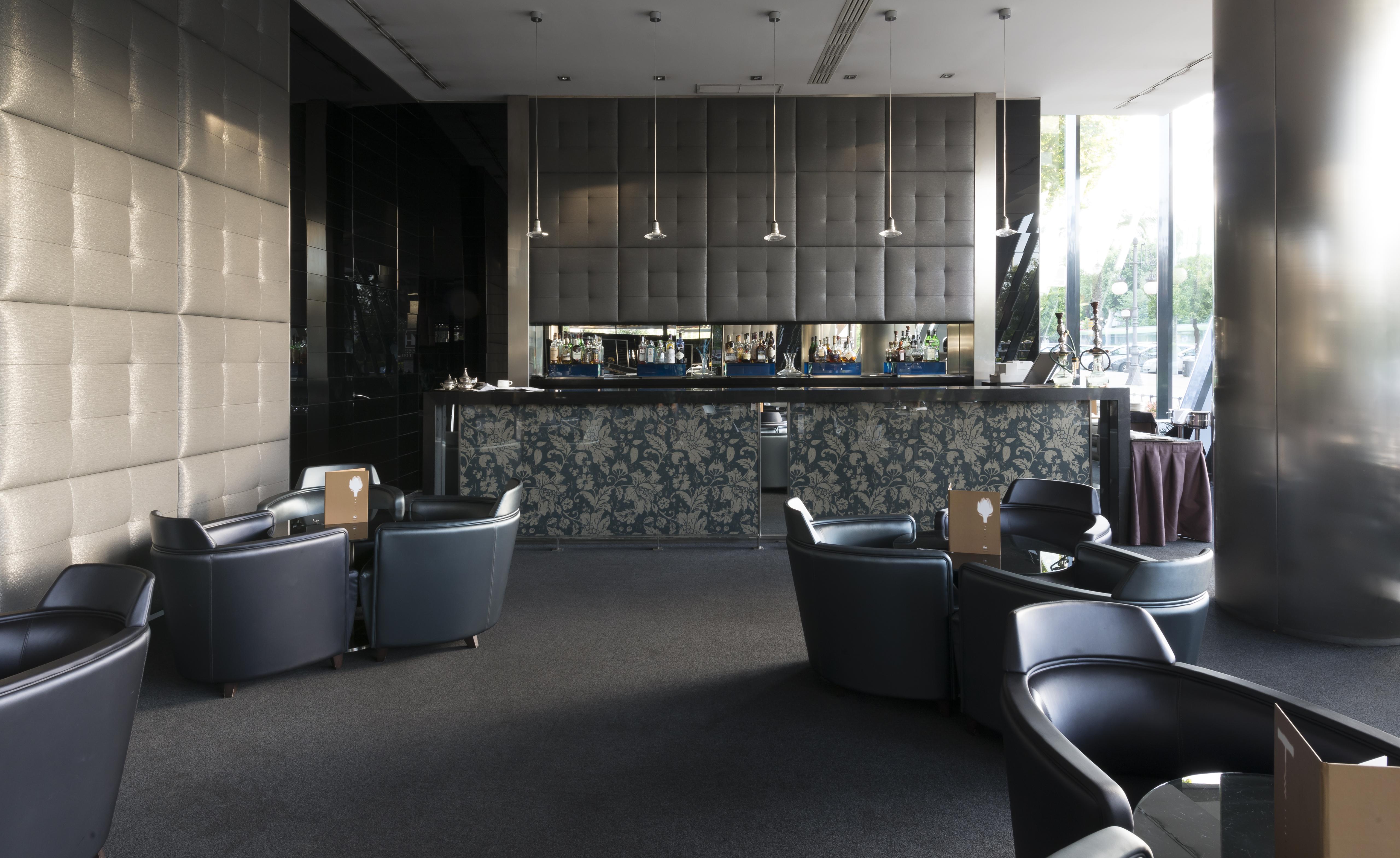 Eurostars Palace Córdoba Exterior foto The photo shows a modern bar or lounge area. In the foreground, there are several comfortable black armchairs arranged around small tables. The background features a sleek bar counter with a dark, textured wall behind it. The bar is illuminated with 