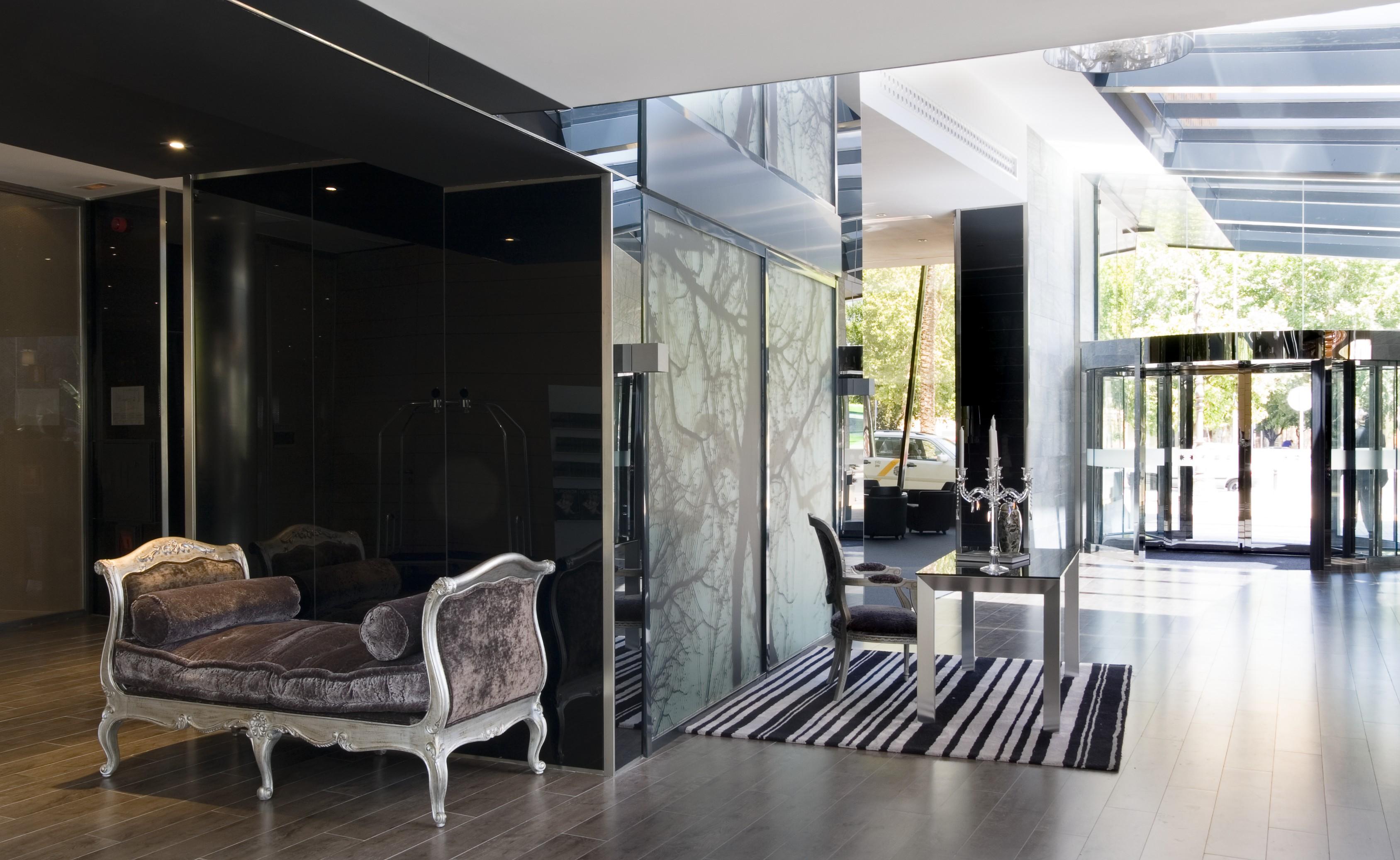 Eurostars Palace Córdoba Exterior foto The photo features a modern and stylish interior space, likely a lobby or reception area. On the left, there is an elegant couch with ornate detailing, set against dark, reflective walls. The floor is wooden and has a sleek design. To the right, ther