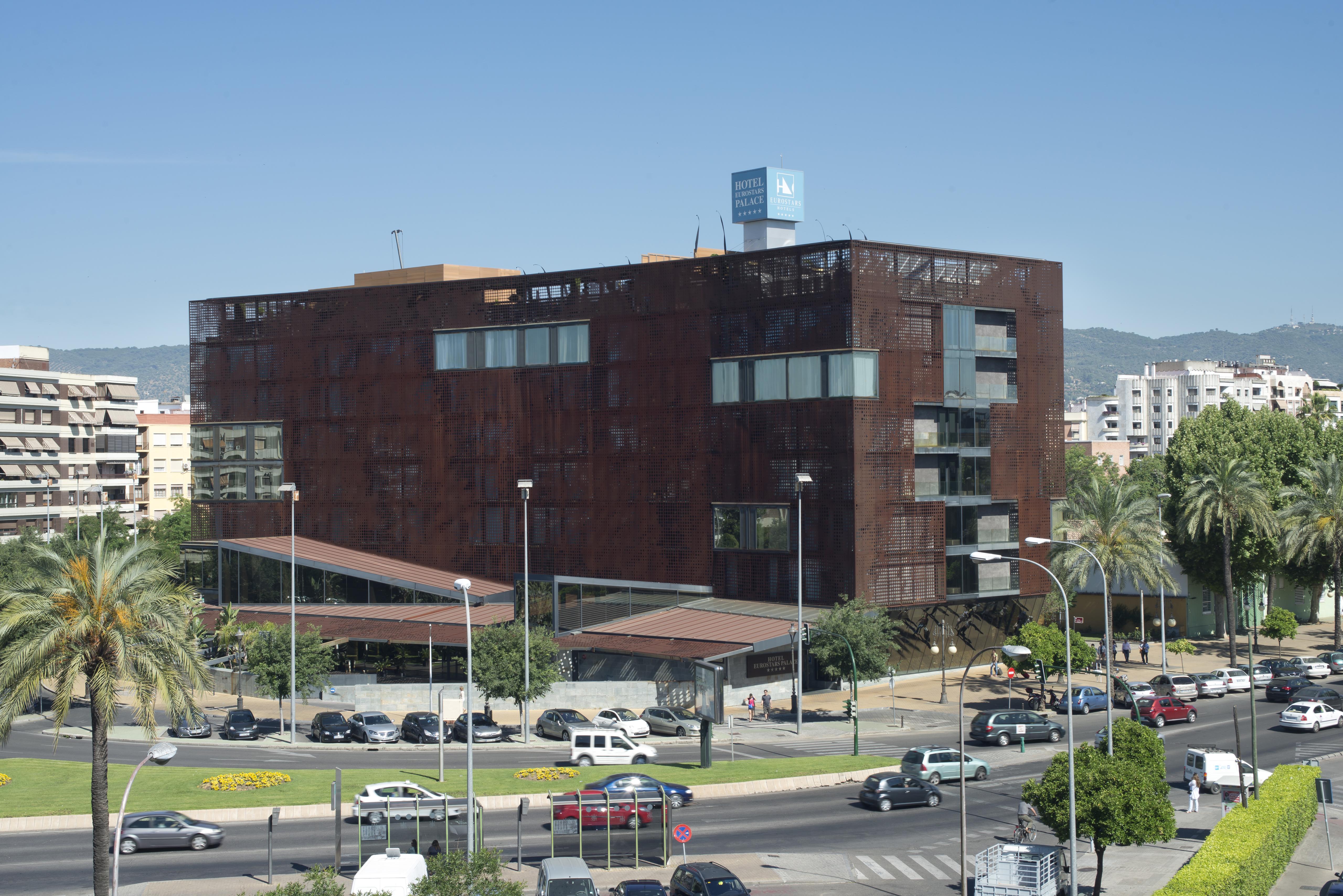 Eurostars Palace Córdoba Exterior foto The photo shows a modern building characterized by a distinctive brown, rust-like exterior, which gives it an industrial appearance. The structure has multiple large windows, allowing for natural light to enter. There are several palm trees surroundi
