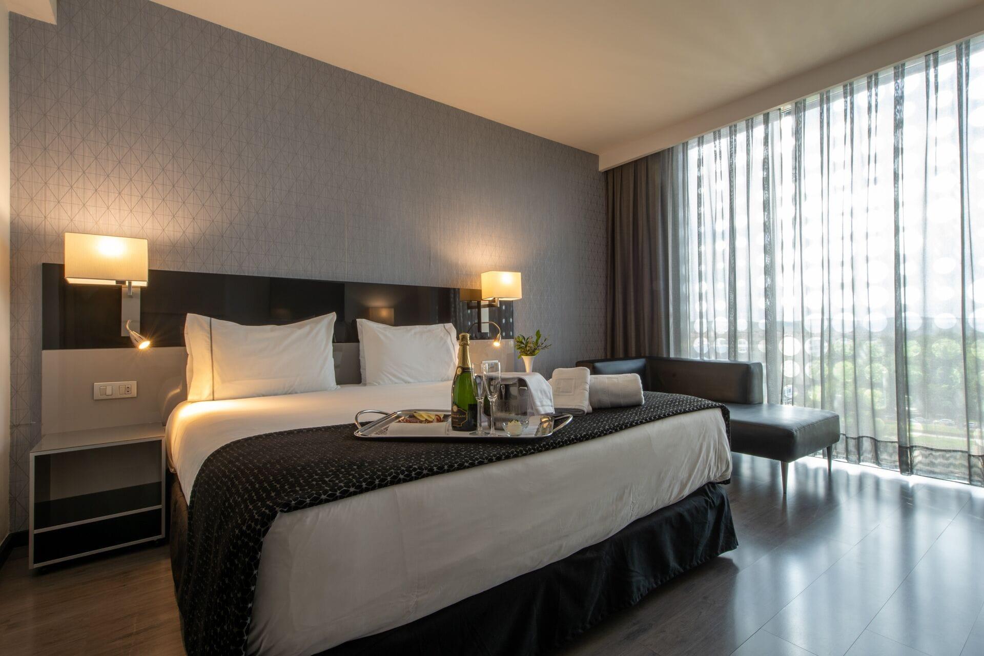 Eurostars Palace Córdoba Exterior foto The photo shows a modern hotel room featuring a large bed with crisp white linens and dark throw blankets. On either side of the bed, there are bedside lamps providing soft lighting. The room has a sleek design with a neutral color palette, including