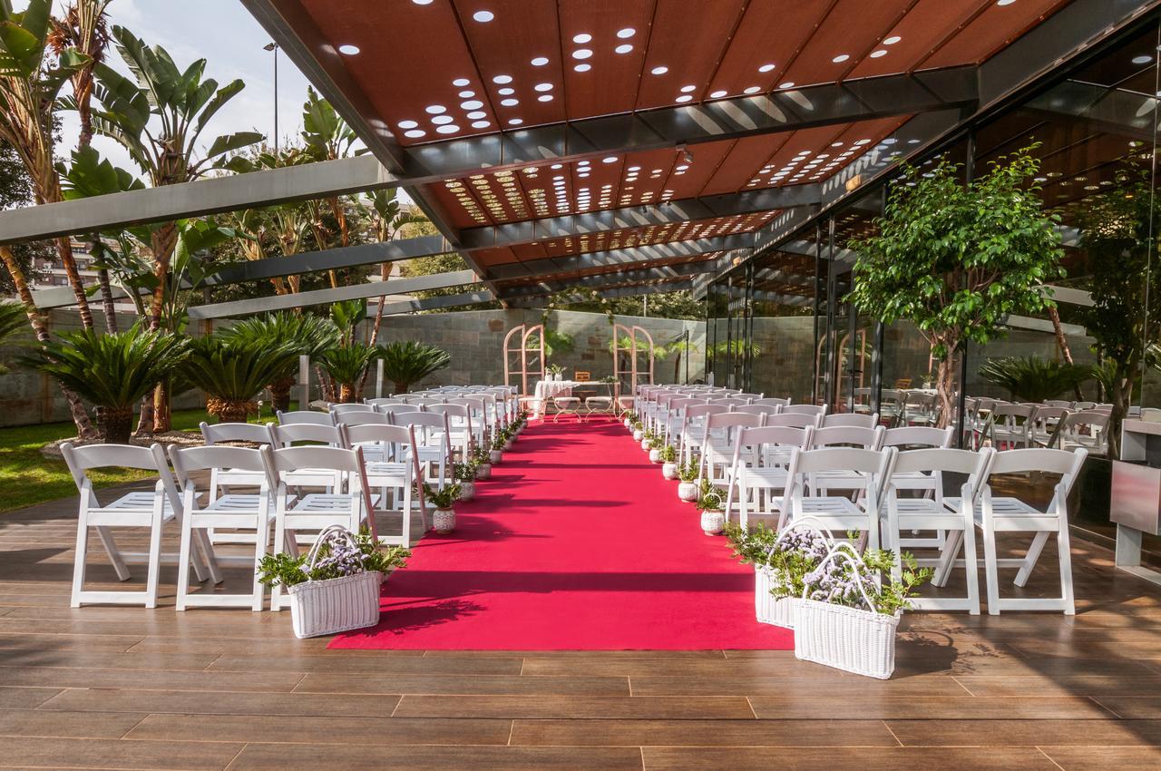 Eurostars Palace Córdoba Exterior foto The image shows an outdoor wedding setup. There are white chairs arranged neatly on either side of a vibrant red carpet that leads to a framed archway. The area is covered by a glass and metal structure, allowing plenty of natural light, while tropic